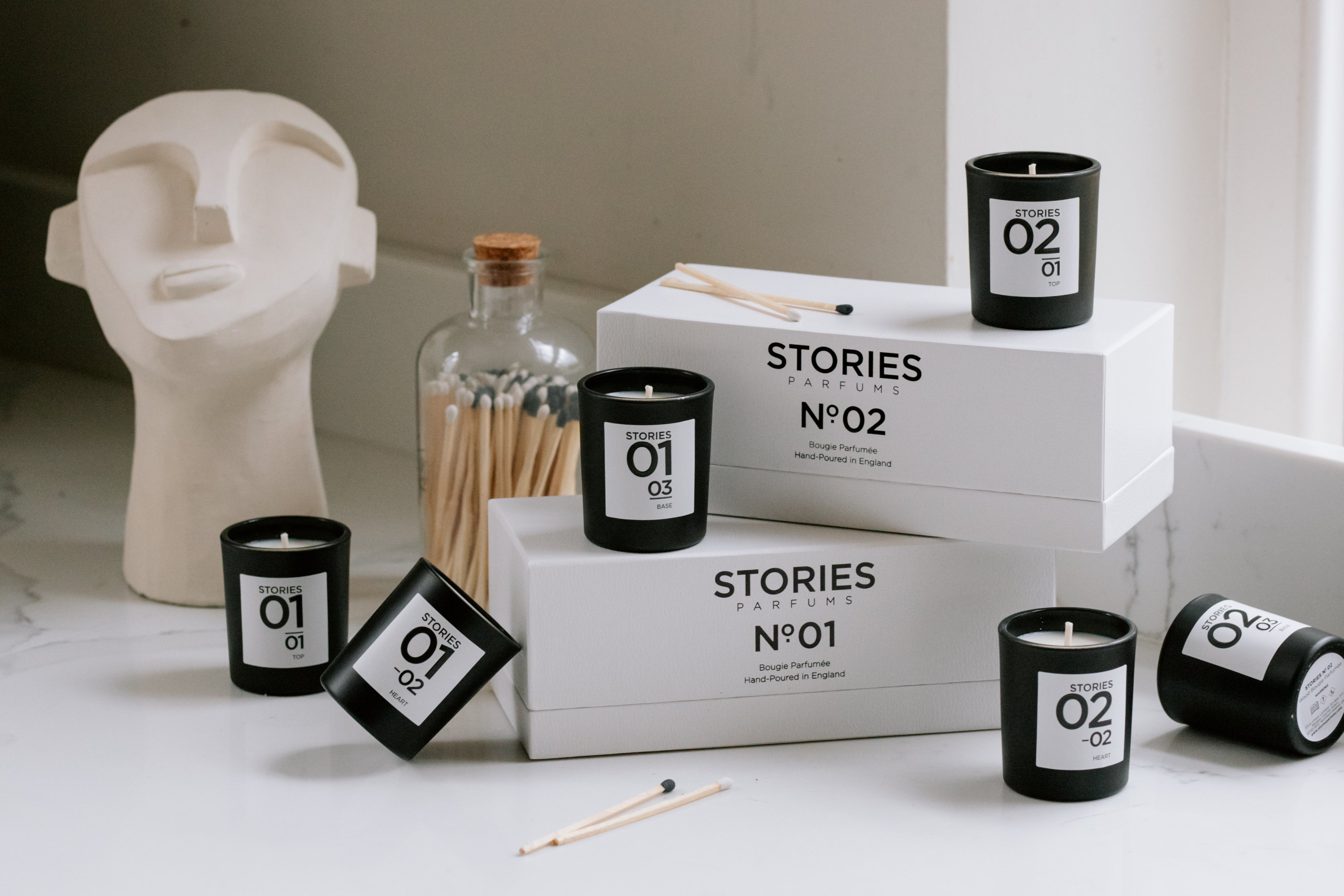small candles in black glass jars arranged around white boxes, next to a jar of matches and decorative white abstract head