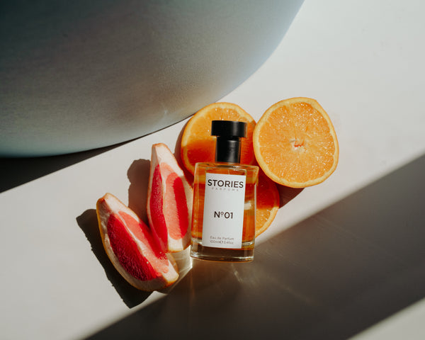 Bottle of STORIES perfume resting on slices of citrus fruit on white shelf