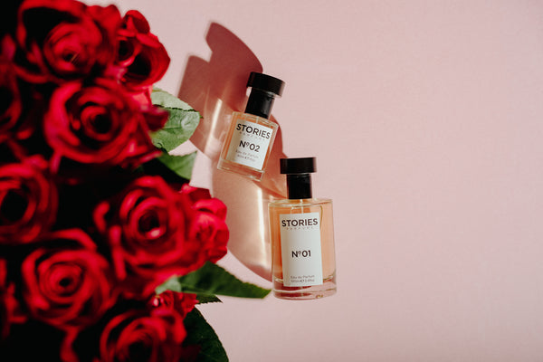 Bottles of STORIES Eau de Parfum lying next to a bunch of red roses on a pale pink background